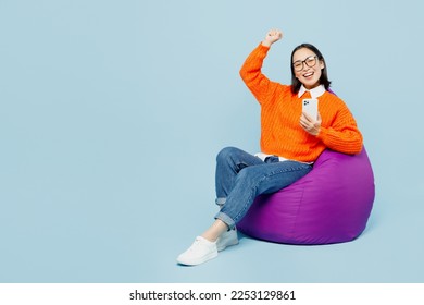 Full body young woman of Asian ethnicity wear orange sweater glasses sit in bag chair hold in hand use mobile cell phone show thumb up do winner gesture isolated on plain pastel blue cyan background - Shutterstock ID 2253129861