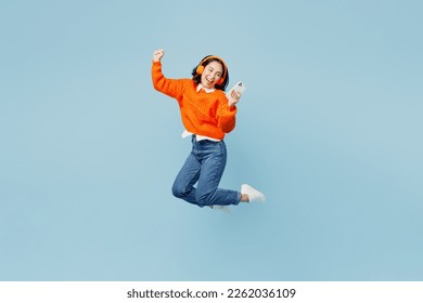 Full body young woman of Asian ethnicity wear orange sweater glasses headphones listen to music raise up hands dance jump high use mobile cell phone isolated on plain pastel light blue cyan background