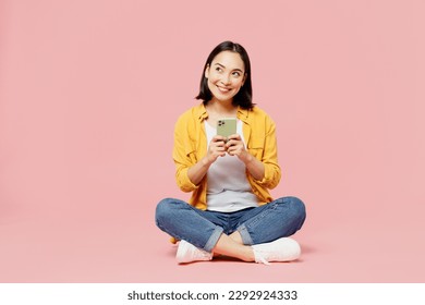 Full body young woman of Asian ethnicity wear yellow shirt white t-shirt sitting hold in hand use mobile cell phone isolated on plain pastel light pink background studio portrait. Lifestyle concept