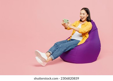 Full body young woman of Asian ethnicity in yellow shirt white t-shirt sit in bag chair use play racing app on mobile cell phone gadget smartphone for pc video games isolated on plain pink background