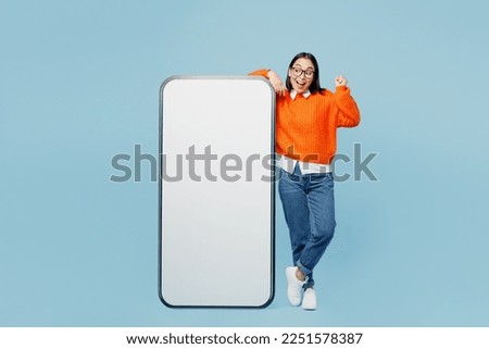 Full body young woman of Asian ethnicity wear orange sweater glasses big huge blank screen mobile cell phone smartphone with area do winner gesture isolated on plain pastel light blue cyan background