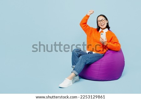 Full body young woman of Asian ethnicity wear orange sweater glasses sit in bag chair hold in hand use mobile cell phone show thumb up do winner gesture isolated on plain pastel blue cyan background