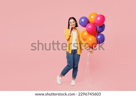 Full body young woman of Asian ethnicity wear yellow shirt white t-shirt hold face bunch of colorful air balloons look camera isolated on plain pastel light pink background studio. Lifestyle concept