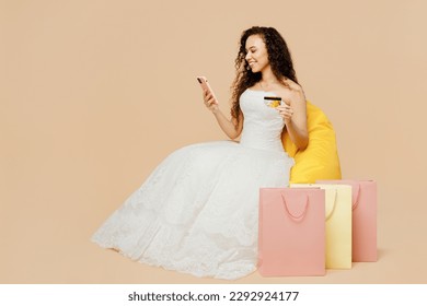 Full body young woman bride wear wedding dress posing sit in bag chair use mobile cell phone credit bank card shopping online isolated on plain beige background studio portrait. Ceremony party concept - Powered by Shutterstock