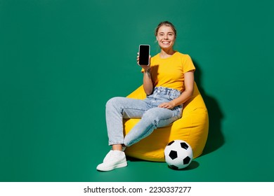 Full body young woman fan wear yellow t-shirt cheer up football sport team sit in bag chair near soccer ball watch tv live stream use mobile cell phone blank screen isolated on dark green background - Powered by Shutterstock