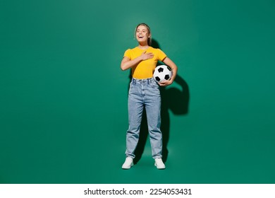 Full body young woman fan wear basic yellow t-shirt cheer up support football sport team hold soccer ball watch tv live stream put hand in heart sing hymn song isolated on dark green background studio