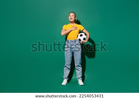 Full body young woman fan wear basic yellow t-shirt cheer up support football sport team hold soccer ball watch tv live stream put hand in heart sing hymn song isolated on dark green background studio