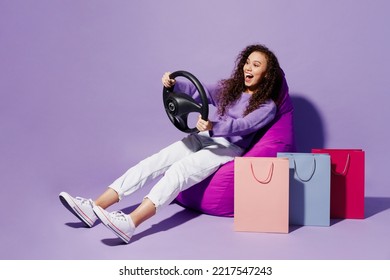 Full Body Young Woman In Pullover Sit In Chair Near Paper Package Bag After Shopping Hold Steering Wheel Driving Car Isolated On Plain Pastel Light Purple Background Black Friday Sale Buy Day Concept