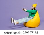 Full body young woman she wear green sweater yellow hat casual clothes sit in bag chair hold in hand play pc game with joystick console isolated on plain pastel light purple background studio portrait