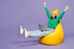 Full Body Young IT Woman She Wear Green Sweater Yellow Hat Casual Clothes Sit In Bag Chair Hold Use Work On Laptop Pc Computer Do Winner Gesture Isolated On Plain Pastel Light Purple Background Studio