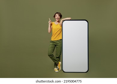 Full Body Young Woman She Wear Yellow T-shirt Near Big Huge Blank Screen Mobile Cell Phone With Workspace Copy Space Mockup Area Point Index Finger Up Isolated On Plain Olive Green Khaki Background.