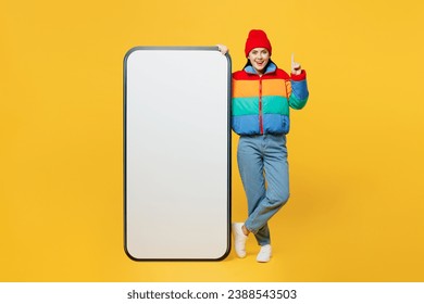 Full body young woman she wears padded windbreaker jacket red hat casual clothes big huge blank screen mobile cell phone smartphone with area point finger up isolated on plain yellow background studio