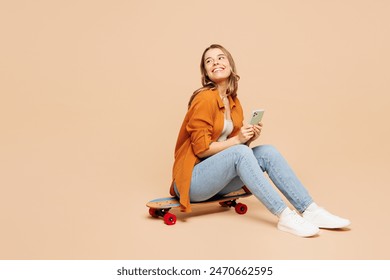 Full body young woman she wear orange shirt casual clothes sits on pennyboard skateboard hold in hand use mobile cell phone isolated on plain pastel light beige background studio. Lifestyle concept - Powered by Shutterstock
