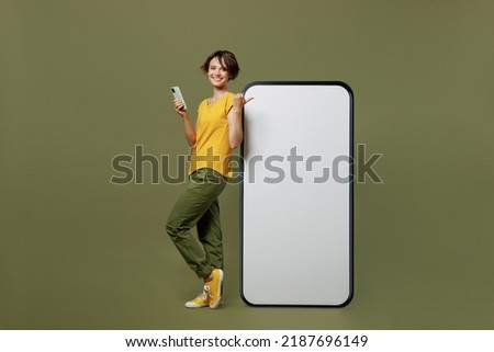 Full body young woman she in yellow t-shirt point finger on big huge blank screen mobile cell phone with workspace copy space mockup area use smartphone isolated on plain olive green khaki background