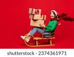 Full body young woman wear green turtleneck Santa hat posing sit ride sled hold present boxes with gift bow isolated on plain red background. Happy New Year 2024 celebration Christmas holiday concept