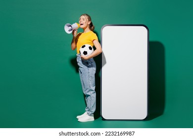 Full body young woman wear yellow t-shirt cheer up support football sport team hold soccer ball watch tv live stream big blank screen mobile cell phone scream in megaphone isolated on green background - Powered by Shutterstock