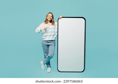Full body young woman wear striped hoody point finger on big huge blank screen mobile cell phone smartphone with workspace copy space mockup area isolated on plain pastel light blue cyan background - Shutterstock ID 2297186063