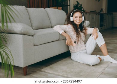 Full body young woman wear casual clothes headphones sit near grey sofa use mobile cell phone listen music stay at home hotel flat rest relax spend free spare time in living room indoor Lounge concept - Shutterstock ID 2367234497