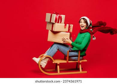 Full body young woman wear green turtleneck Santa hat posing sit ride sled hold present boxes with gift bow isolated on plain red background. Happy New Year 2024 celebration Christmas holiday concept