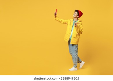 Mujer joven de cuerpo entero usa impermeable ropa interior impermeable sombrero rojo para selfie en el show de teléfono celular v-sign aislado en el fondo amarillo plano. Concepto de la estación meteorológica de otoño húmedo al aire libre