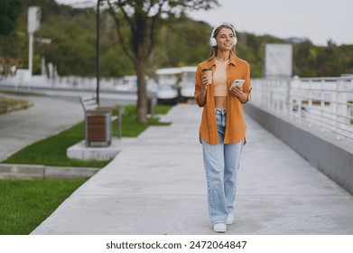 Full body young woman wear orange shirt casual clothes listen music in headphones use mobile cell phone drink coffee rest relax walk in spring city park outside in summer day. Urban lifestyle concept - Powered by Shutterstock