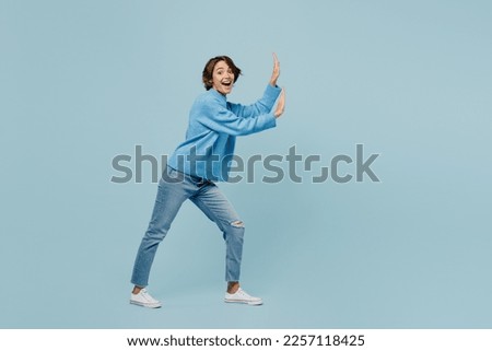 Full body young woman wear knitted sweater look camera push invisible object with copy space area mock up isolated on plain pastel light blue cyan background studio portrait. People lifestyle concept