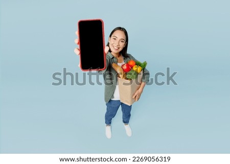 Full body young woman wear casual clothes hold brown paper bag with food products close up blank screen mobile cell phone isolated on plain blue background. Delivery service from shop or restaurant