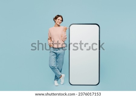Full body young woman wear beige knitted sweater casual clothes point index finger on big huge blank screen mobile cell phone smartphone with workspace area isolated on plain pastel blue background