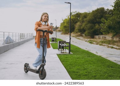 Full body young woman wearing orange shirt casual clothes headphones riding e-scooter using mobile cell phone walking rest relax in spring green city park outdoors on nature. Urban lifestyle concept