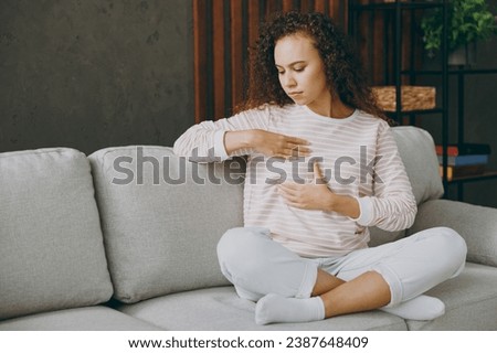 Full body young woman wearing casual clothes sits on grey sofa couch hold touch breast check health stay at home hotel flat rest relax spend free spare time in living room indoor. Healthcare concept