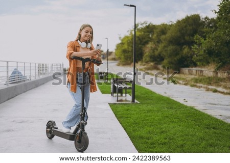 Full body young woman wearing orange shirt casual clothes headphones riding e-scooter using mobile cell phone walking rest relax in spring green city park outdoors on nature. Urban lifestyle concept