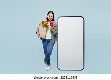 Full body young woman wears casual clothes hold bag with food products big huge blank screen area use mobile cell phone isolated on plain blue background studio portrait. Delivery service from shop - Powered by Shutterstock