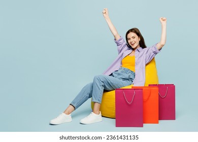 Full body young woman wears casual clothes sit in bag chair near paper package bags after shopping do winner gesture isolated on plain pastel blue cyan background. Black Friday sale buy day concept