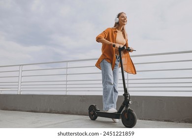 Full body young woman wears orange shirt casual clothes riding e-scooter transport listen music in headphones walking rest relax in spring green city park outdoors on nature. Urban lifestyle concept - Powered by Shutterstock
