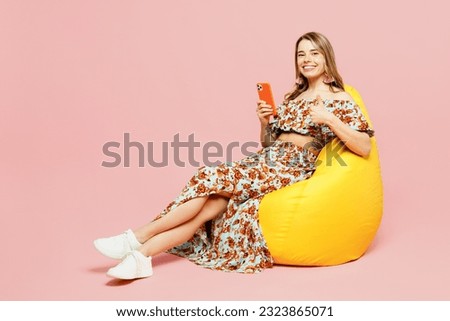 Full body young woman wears summer casual clothes sit in bag chair hold in hand use mobile cell phone show thumb up isolated on plain pastel light pink background studio portrait. Lifestyle concept