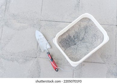 Full Bucket Of Polymeric Paver Sand With Trowel On Top Of Gray Concrete Patio Stone Paver Walkway. This Material To Fill Joint Between Pavers To Firm And Prevent Weeds From Growing Up Between