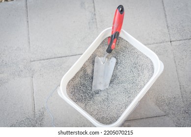 Full Bucket Of Polymeric Paver Sand With Trowel On Top Of Gray Concrete Patio Stone Paver Walkway. This Material To Fill Joint Between Pavers To Firm And Prevent Weeds From Growing Up Between