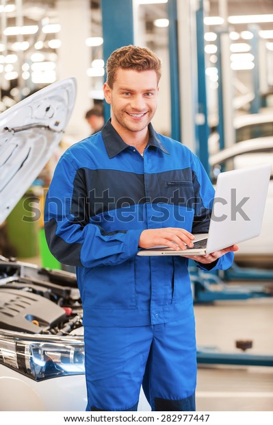 Full of confidence and expertise. Confident\
young man working on laptop and smiling while standing in workshop\
with car in the background