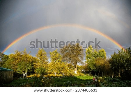 The full double rainbow scenery above the courtyard garden. Saturated rainbow and pale reslection above it. Natural optical phenomenon on the sky. Grey gloomy sky after rain. Sun light on tree tops.