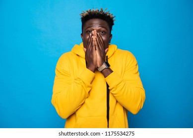 Full Of Emotion. Waist Up Portrait Of Young Man In Yellow Hoodie Hiding Lips And Nose Under Arms. He Looking At Camera With Wide Opened Eyes And Raised Eyebrows As If Found Out Happy News