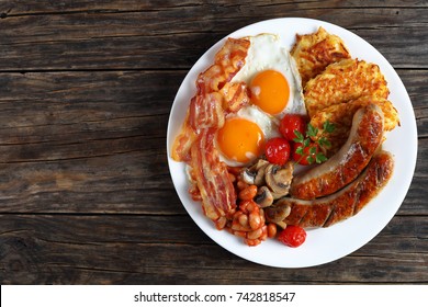 full english breakfast - bean, hash brown, fried eggs sunny side up, bacon slices, sausages, tomatoes, mushrooms on white plate on dark wooden table, view from above 