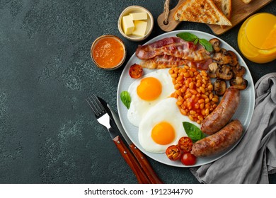 Full English breakfast on a plate with fried eggs, sausages, bacon, beans, toasts and coffee on dark stone background. With copy space. Top view.