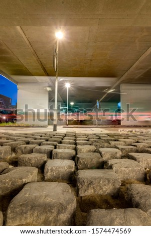 Full focus portrait size background picture. Urban night scene under concrete flyover bridge with square boulder rocks in sand, pedestrian crossing, street lights and trail lights from fast traffic