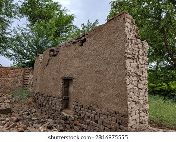 Full frame view of an old mud and red brick wall standing amidst green trees. Wide angle photography capturing abandoned village ruins, weathered clay plaster, and rustic Asian architecture outdoors in daylight.