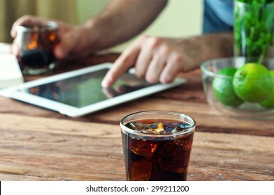 Full Glass Beaker Of Cola On A Wooden Table. The Background Male Hand Click On The Tablet Screen. The Concept Of Work Outside The Office. Copy Space. Close-up. Free Space For Text. View From Above