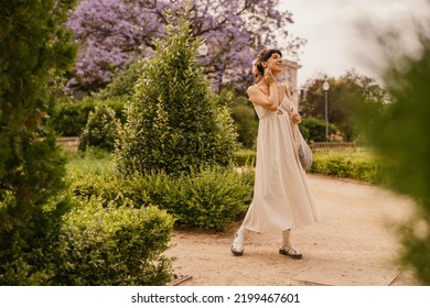 In full growth, pretty young caucasian girl walks in city park summer. Brunette examines nature, wears sundress. Rest time concept - Powered by Shutterstock