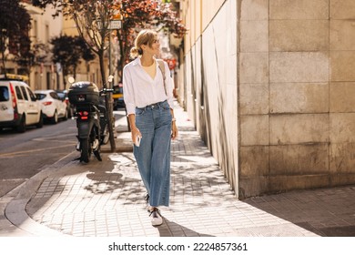 In full growth, pretty young caucasian girl walks around city center in warm weather. Fair-haired lady wears white shirt with jeans on street. Happy weekend concept. - Powered by Shutterstock