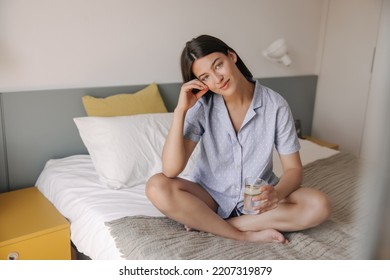 In Full Growth Young Caucasian Girl Wears Pajamas Sitting On Bed. Brunette Hair Woman With Glass Of Water In Morning. Concept Home Comfort.