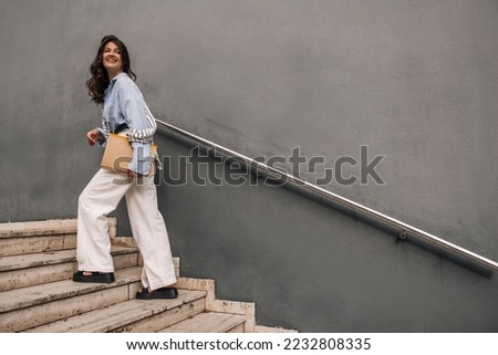 In full growth, young caucasian woman with documents in hands climbs stairs going to work. Brunette wears blue shirt, white trousers and sweatshirt. Business lady concept
