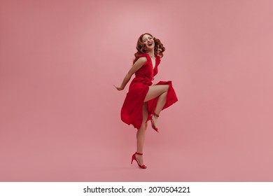 In full growth young red-haired woman dances vigorously for photo in studio. Beauty in red dress with slit and bare leg in high-heeled shoes. Leisure concept of youth, active lifestyle